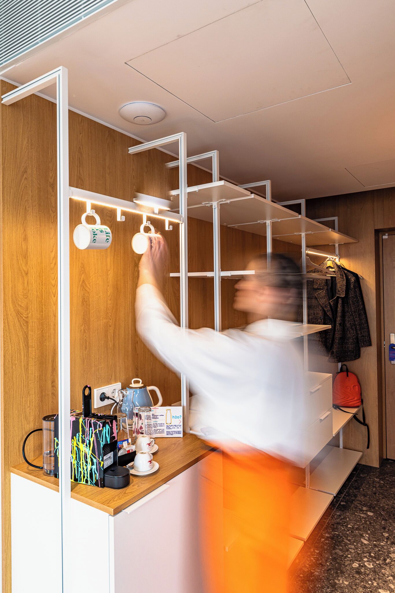 Una habitación de hotel moderna con una pared de madera, un estante blanco con tazas, una cafetera y un bolso rojo.
