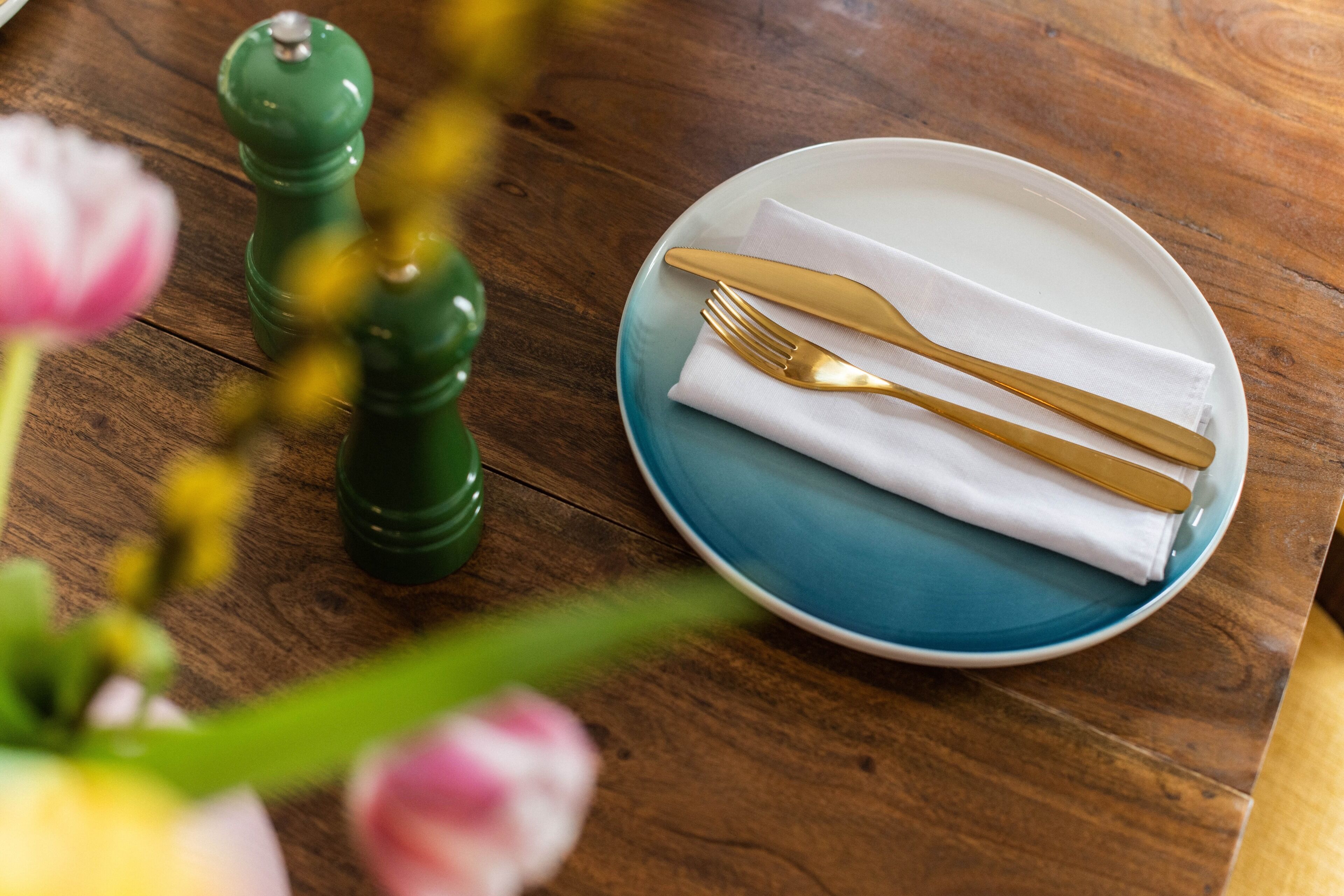 A wooden table with a blue plate, white napkin, and gold cutlery.