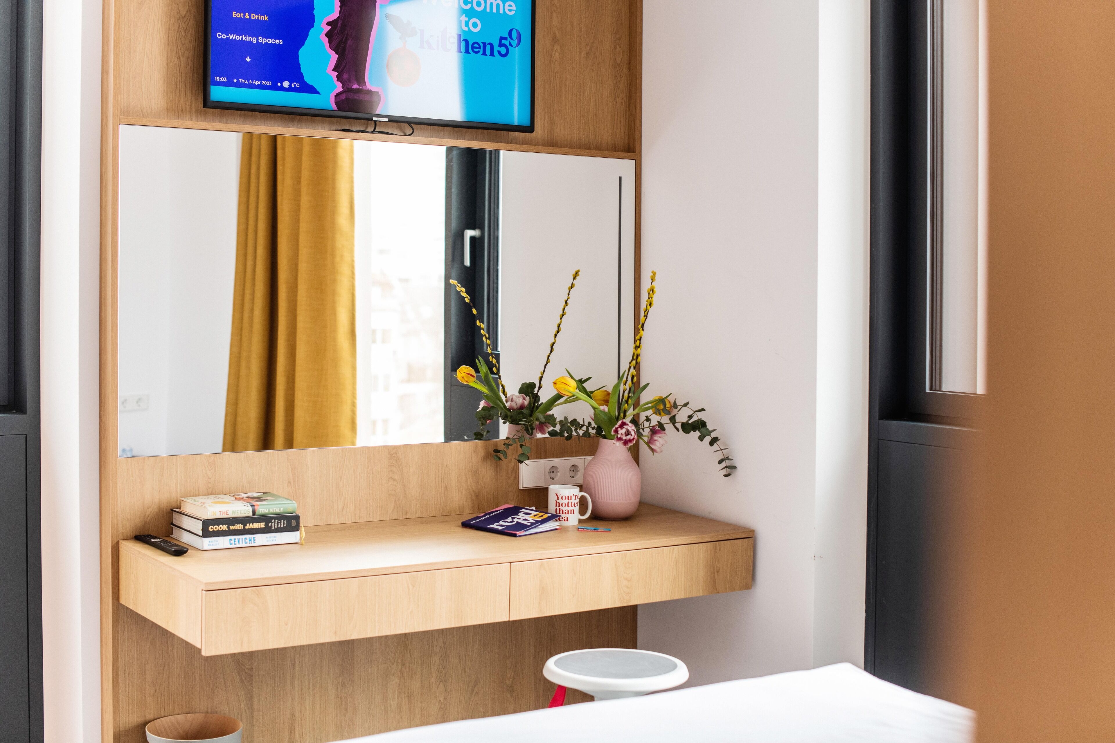 Una habitación de hotel moderna con un escritorio de madera, un espejo, un jarrón con flores y un televisor mostrando un mensaje de bienvenida.