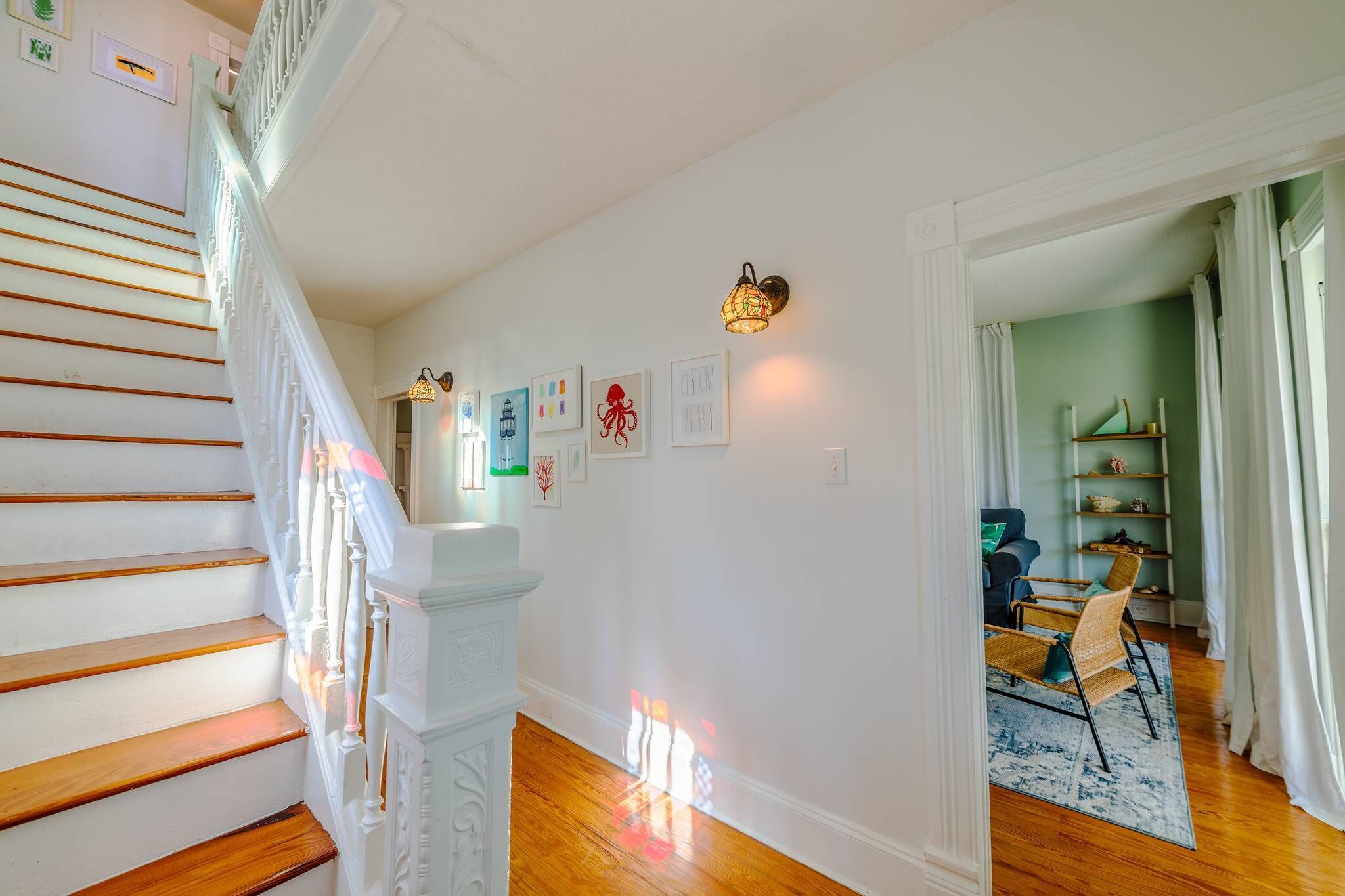 A staircase with white railings and wooden steps, a wall with framed artwork, and a room with a blue rug and wicker chairs.