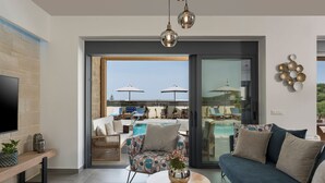 A modern living room with a sofa, a coffee table, and a TV on a brick wall. There is a sliding glass door leading to a pool area with umbrellas and lounge chairs. - Perfect location, private pool, hot tub and gym! (Chania)