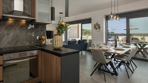 A modern kitchen with a built-in oven, a dining area with a wooden table and chairs, and large windows offering a view of the outdoors. - Perfect location, private pool, hot tub and gym! (Chania)