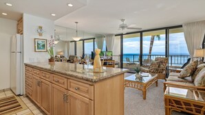 Una cucina moderna con mobili in legno e un'isola centrale, una zona living con divano e poltrone, e ampie finestre che si affacciano sull'oceano.