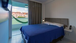 A bedroom with a bed, a nightstand, a lamp, a window with a view of buildings, and a curtain. - Villas do Atlântico próximo ao mar (Ipojuca)