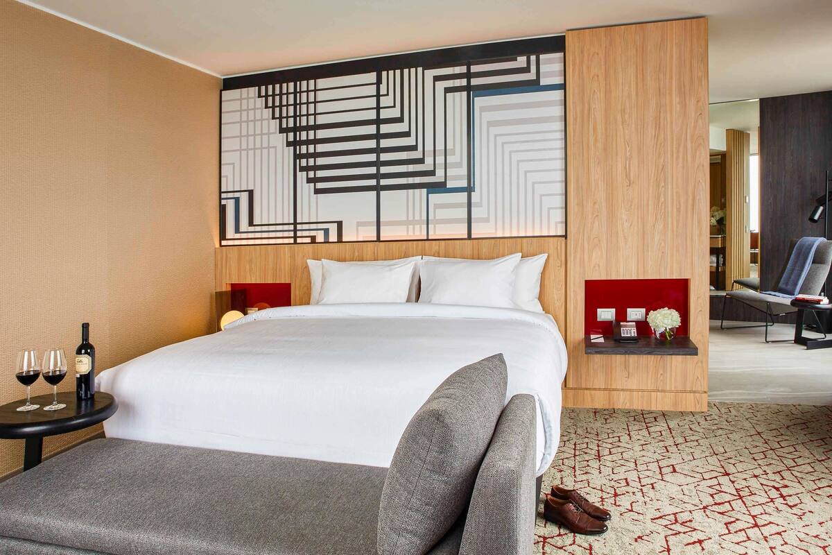 A modern hotel room with a large bed, a small table with wine glasses, a chair, and a decorative wall piece.