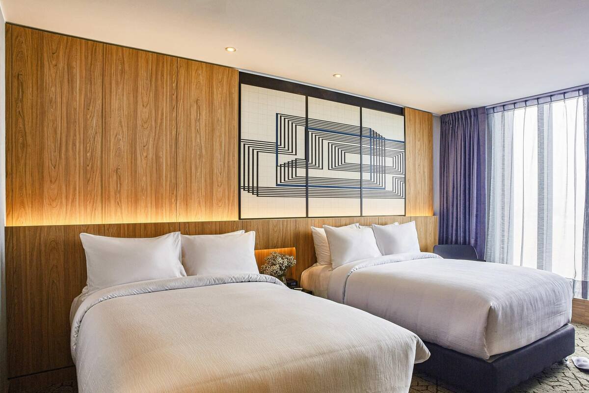 A hotel room with two beds, a wooden accent wall, a geometric art piece, and a view of the outdoors.