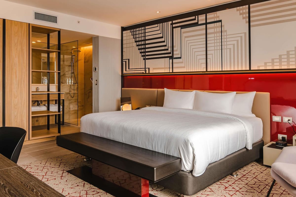 A modern hotel room with a large bed, a desk, a chair, a bench, and a walk-in closet.