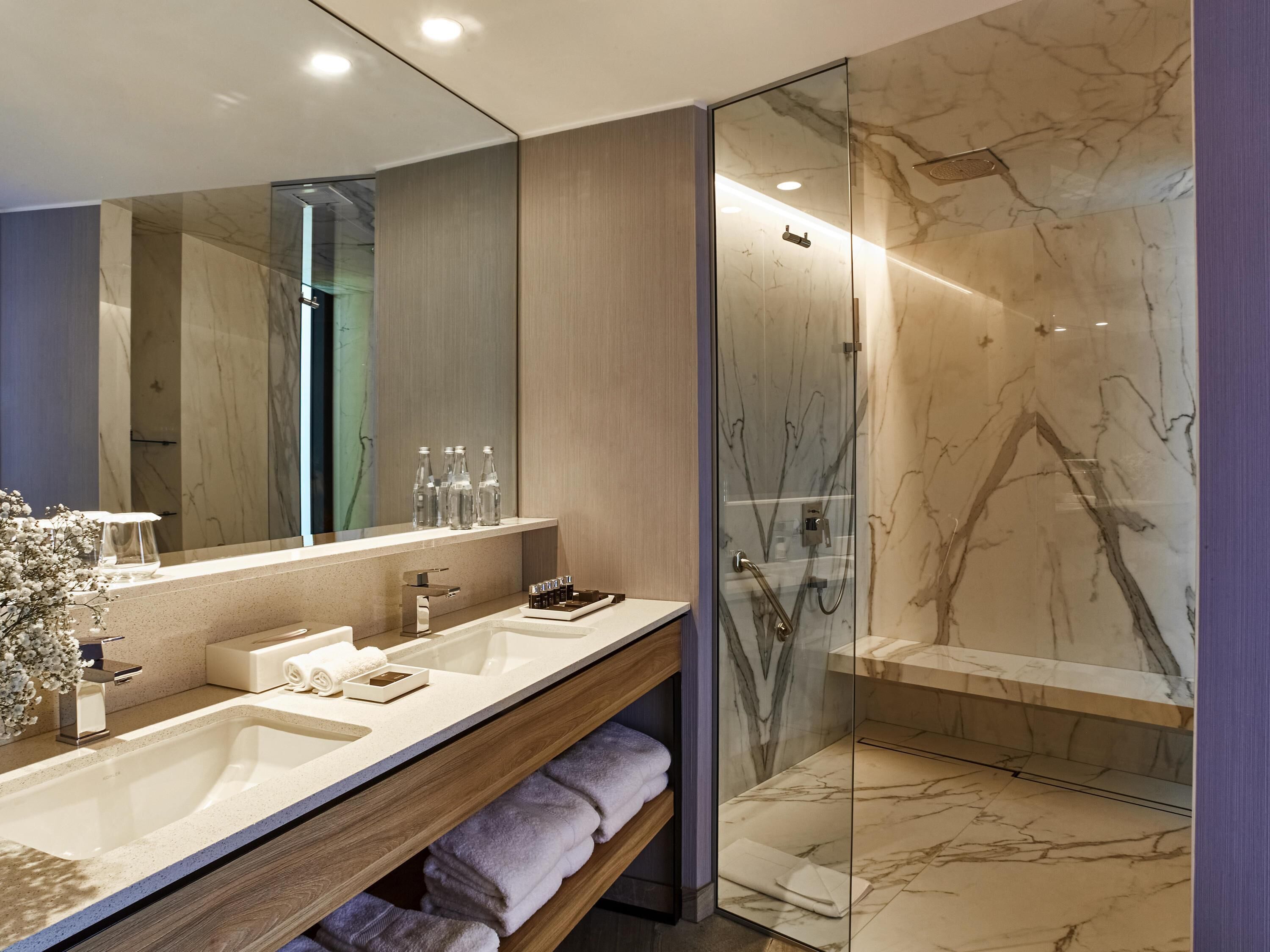A modern bathroom with two sinks, a large mirror, a glass shower enclosure, and marble flooring.