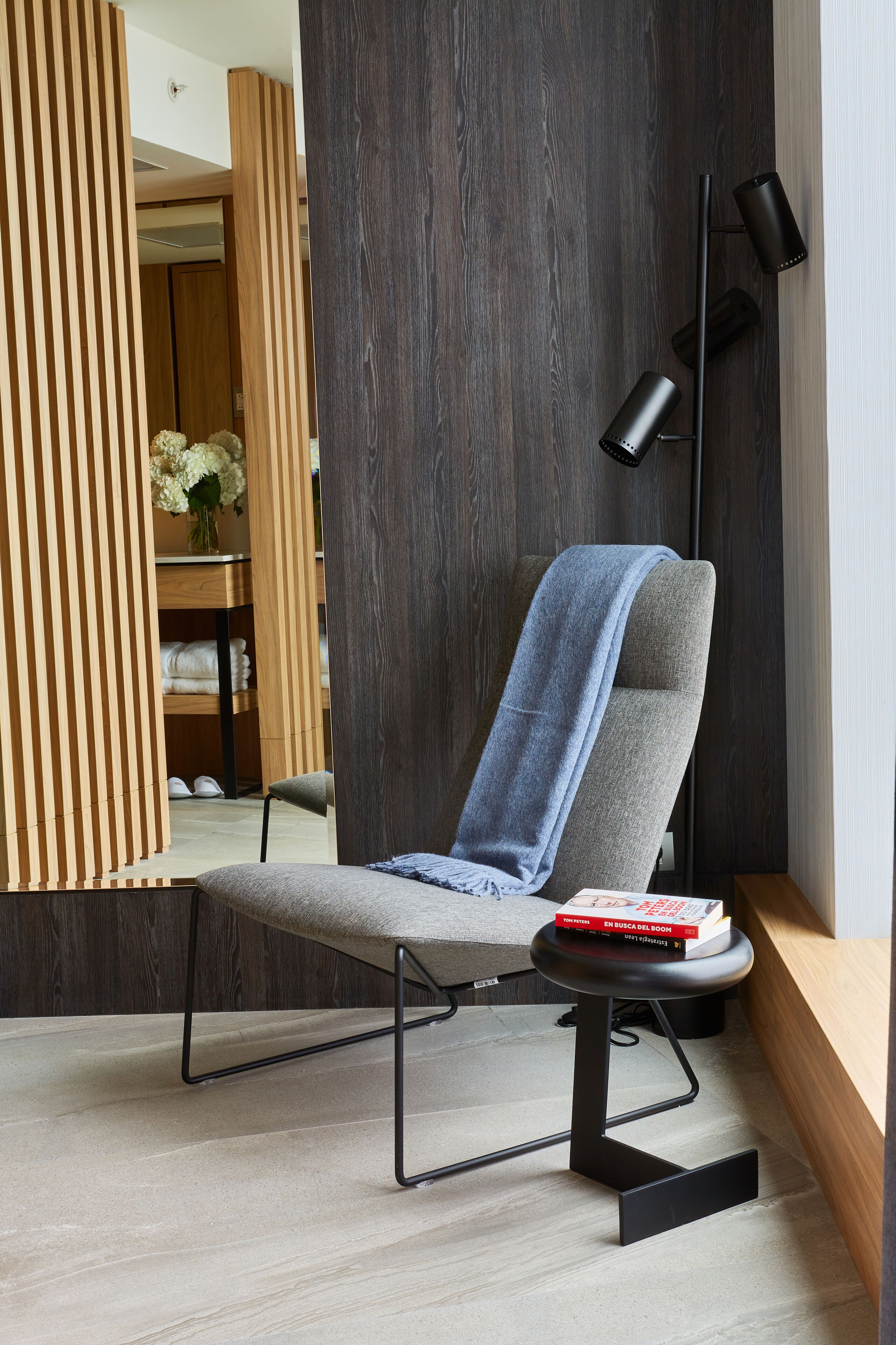 A modern lounge chair with a blue towel, a book, and a black floor lamp in a contemporary room with wooden paneling and a mirror.
