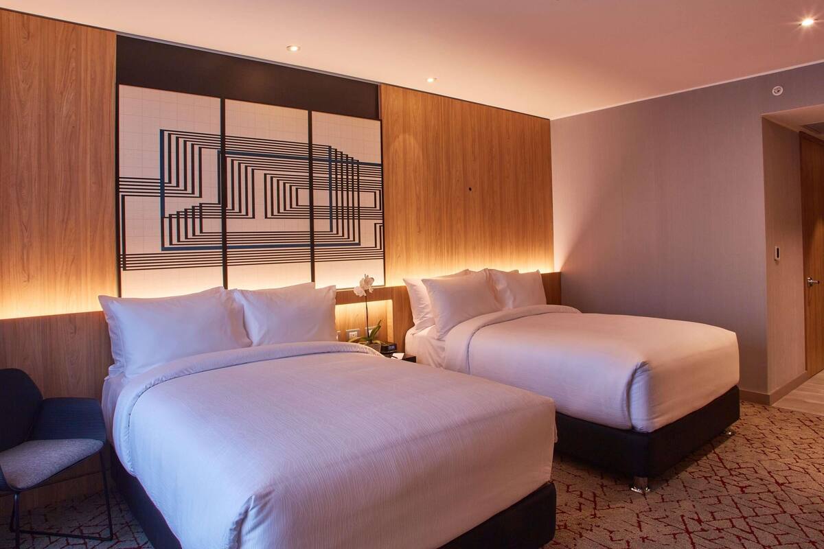 A hotel room with two beds, a wooden headboard, a geometric wall art, and a small desk with a chair.