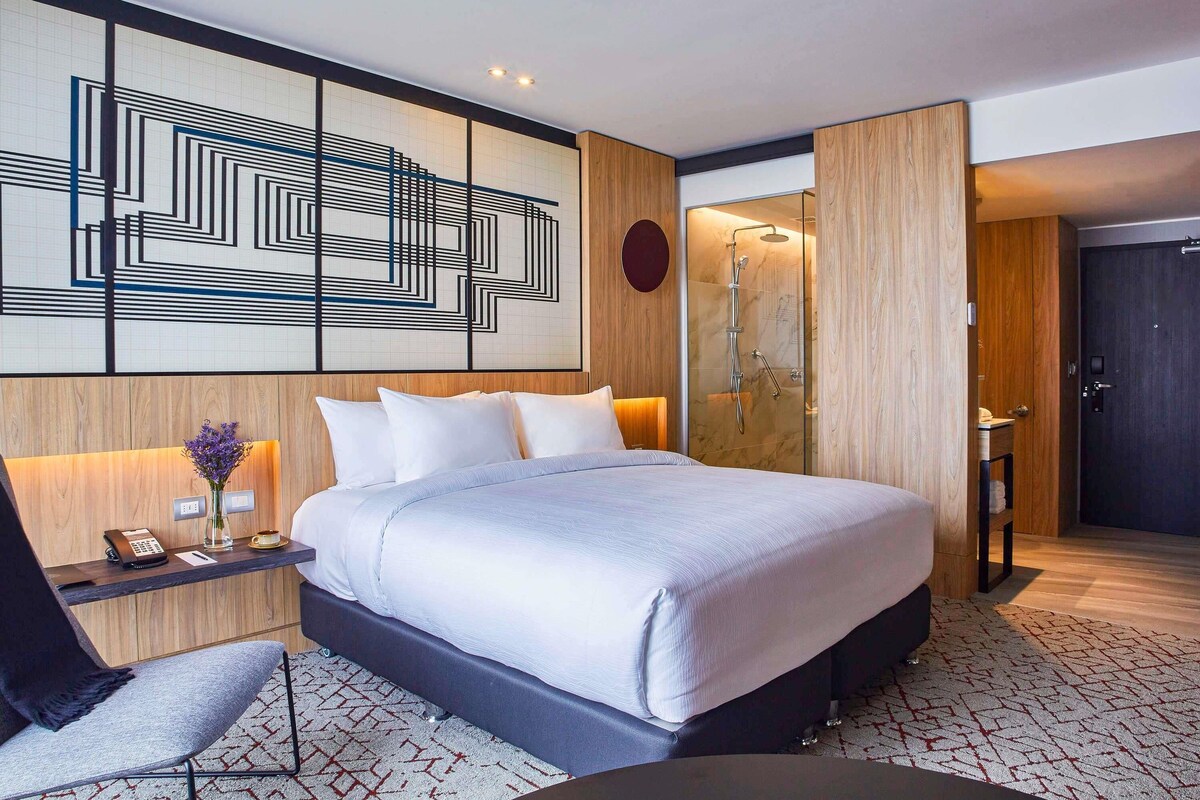 Una habitación de hotel moderna con una cama grande, una mesita de noche con teléfono y jarra, una zona de ducha y una puerta oscura.