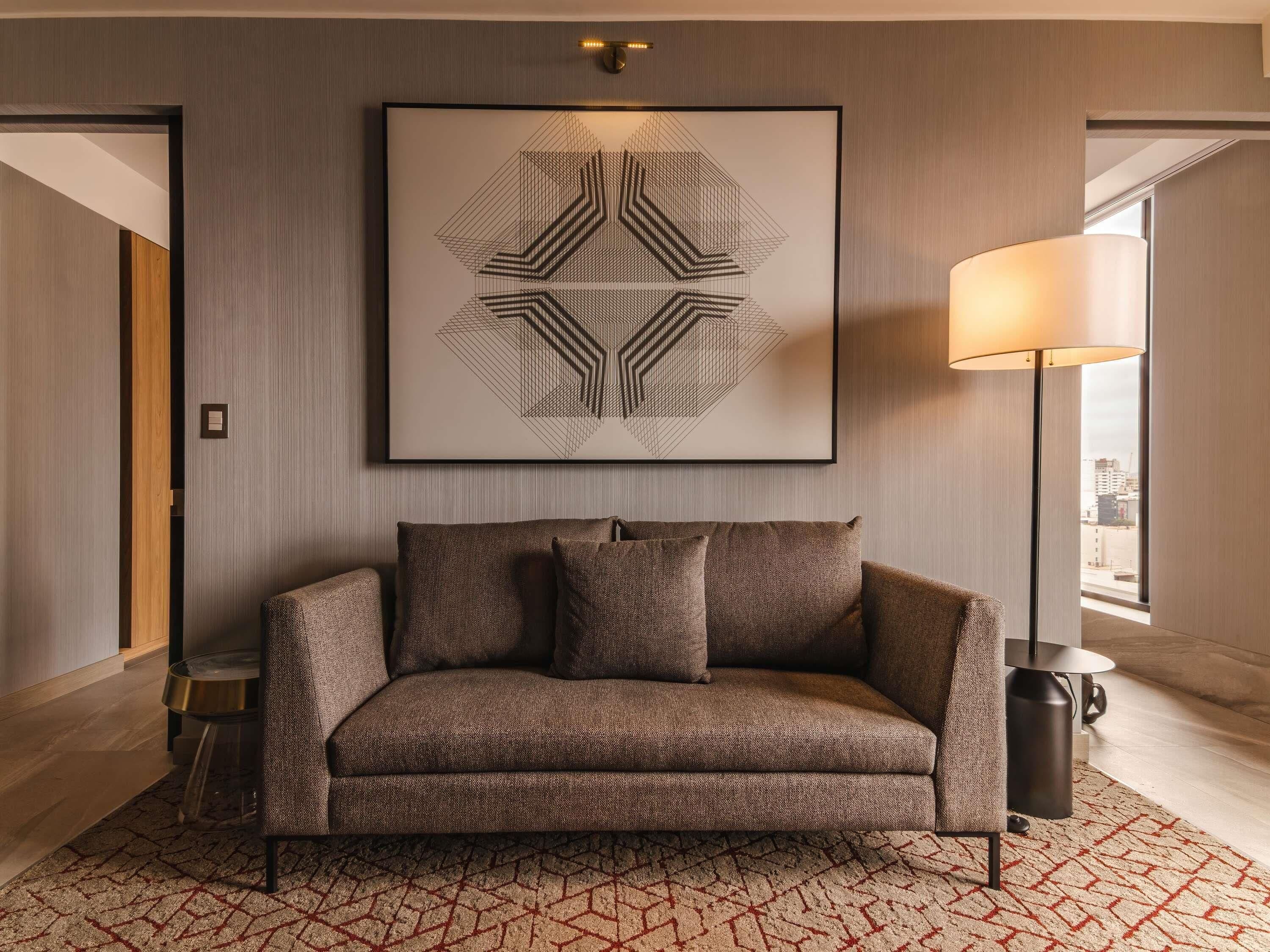 Una habitación de hotel moderna con sofá, una obra de arte geométrica en la pared, una lámpara de pie y vistas a través de una ventana.