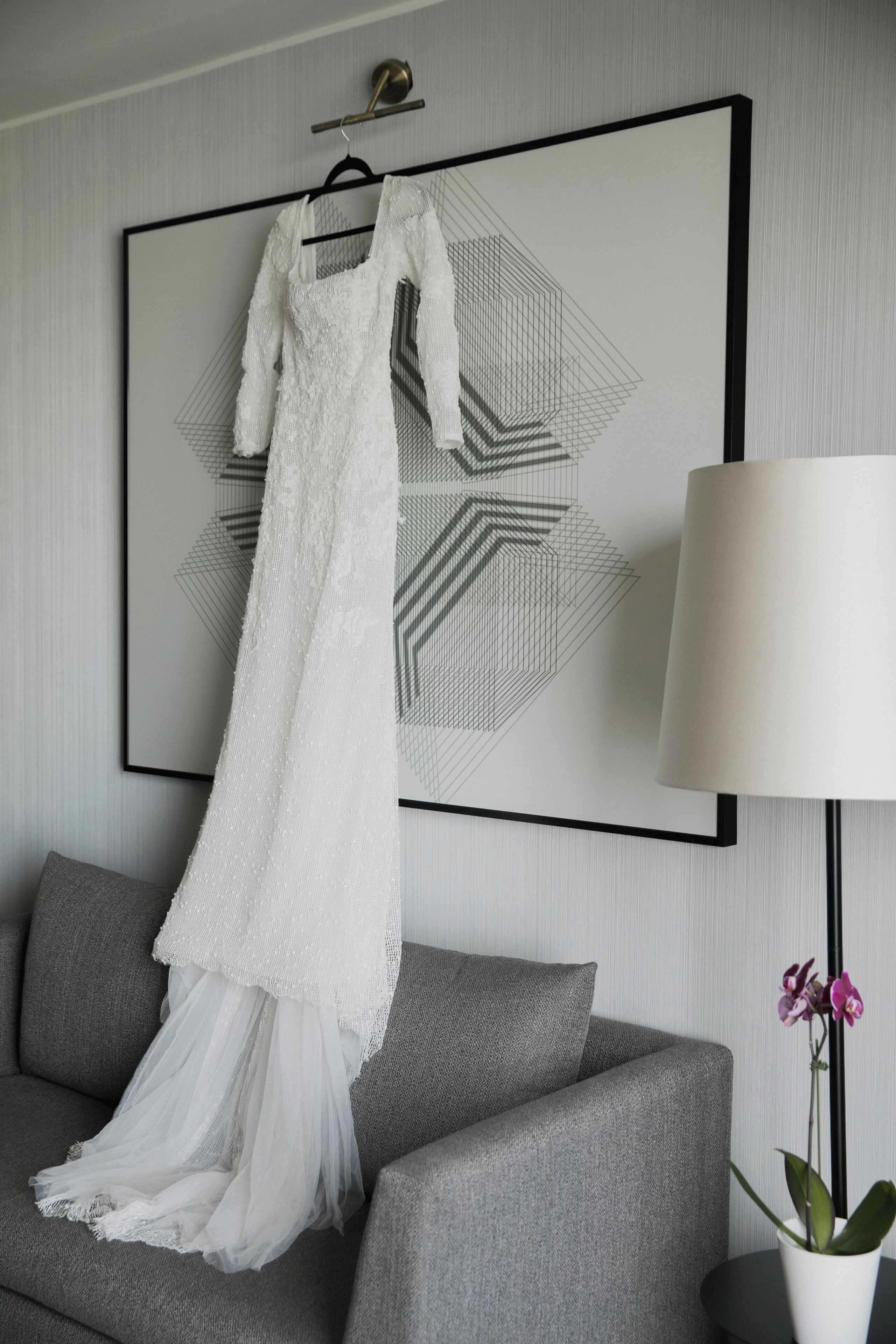 Un vestido de novia blanco colgado en una pared, un sofá gris y una orquídea en maceta.