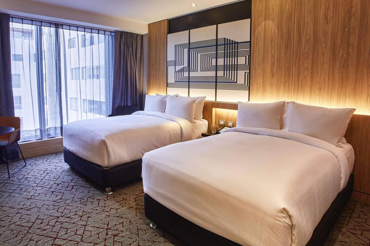 A hotel room with two beds, a large window with curtains, a wall with a geometric design, and a carpeted floor.