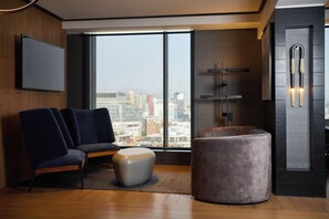 A modern living room with a city view, a flat-screen TV, a fireplace, and a seating area with a round ottoman and two chairs.