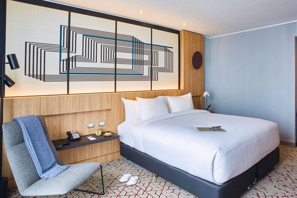 A modern hotel room with a large bed, a desk with a phone and a cup, a chair, and a decorative wall piece.
