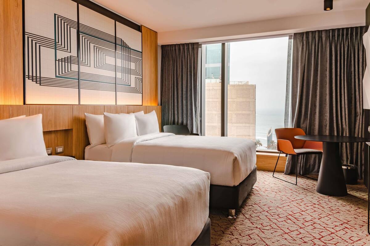 A hotel room with two beds, a desk, and a view of the cityscape.