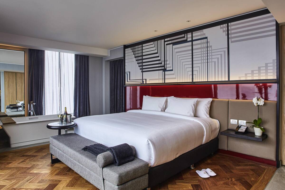 A modern hotel room with a large bed, a bedside table, a bench, a mirror, and a decorative wall panel.