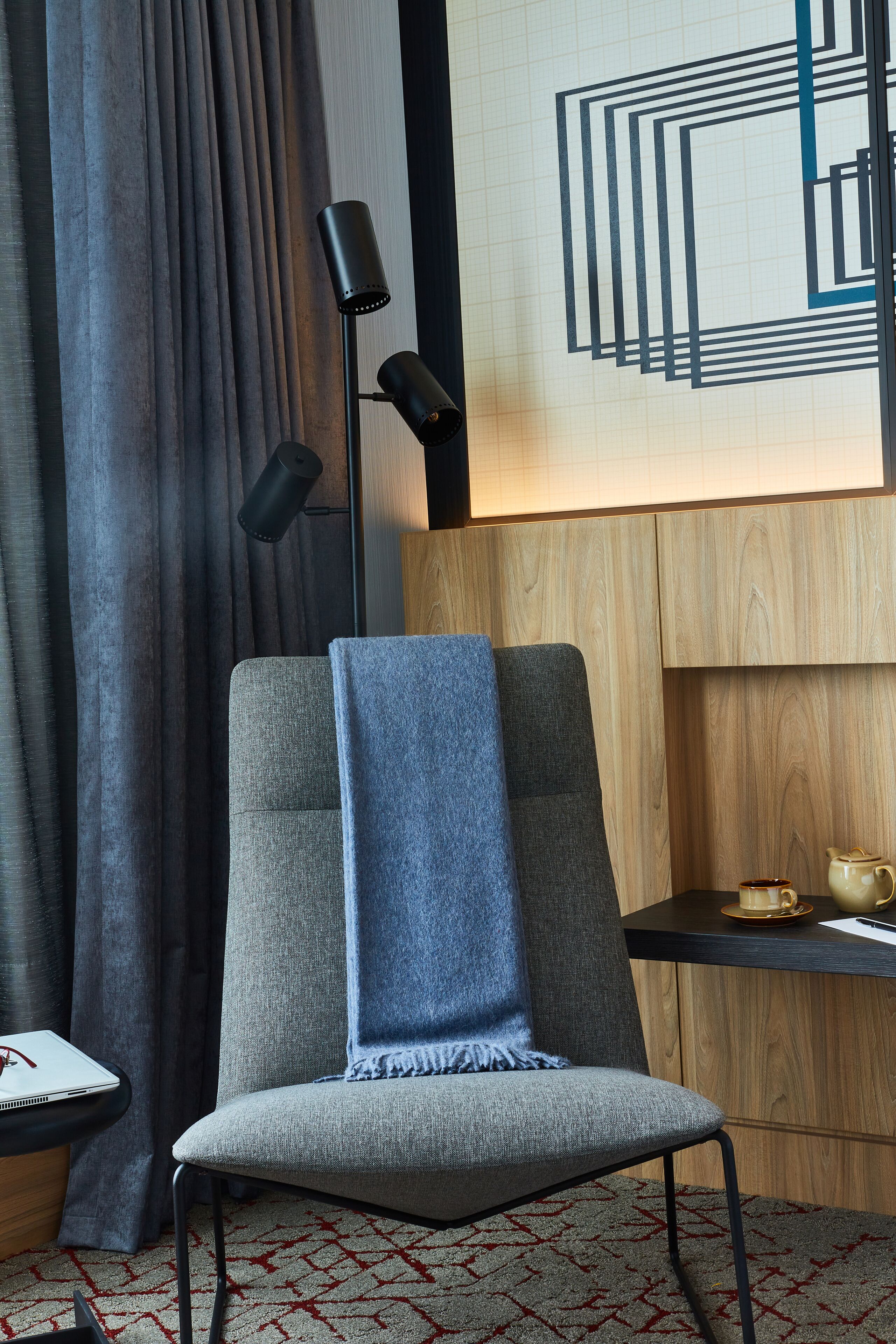 Une pièce moderne avec un fauteuil à revêtement gris, un lampadaire noir, un bureau en bois avec un ordinateur portable et une théière, ainsi qu’un tapis à motifs.