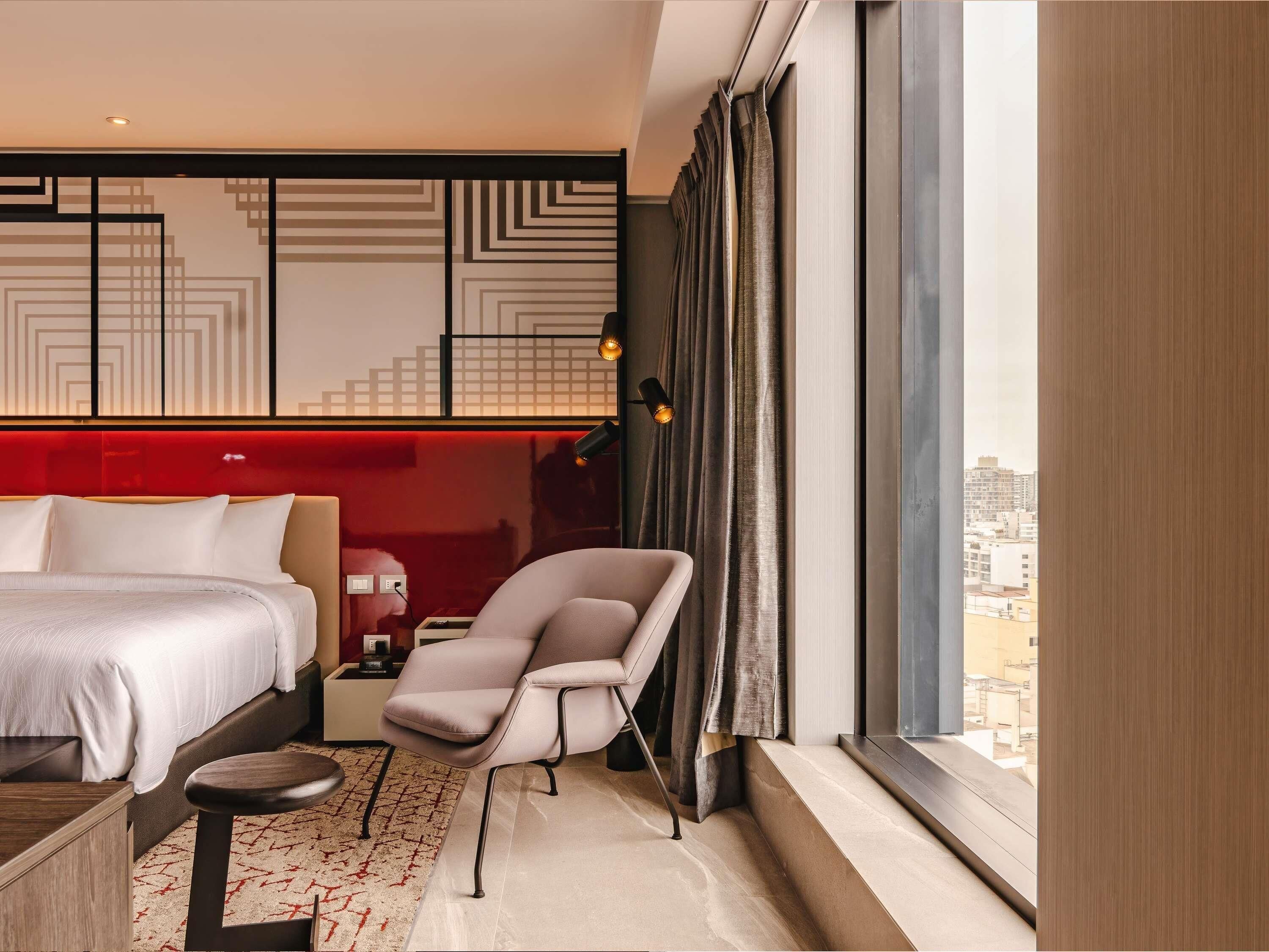 A modern hotel room with a bed, a chair, a desk, and a large window with a city view.
