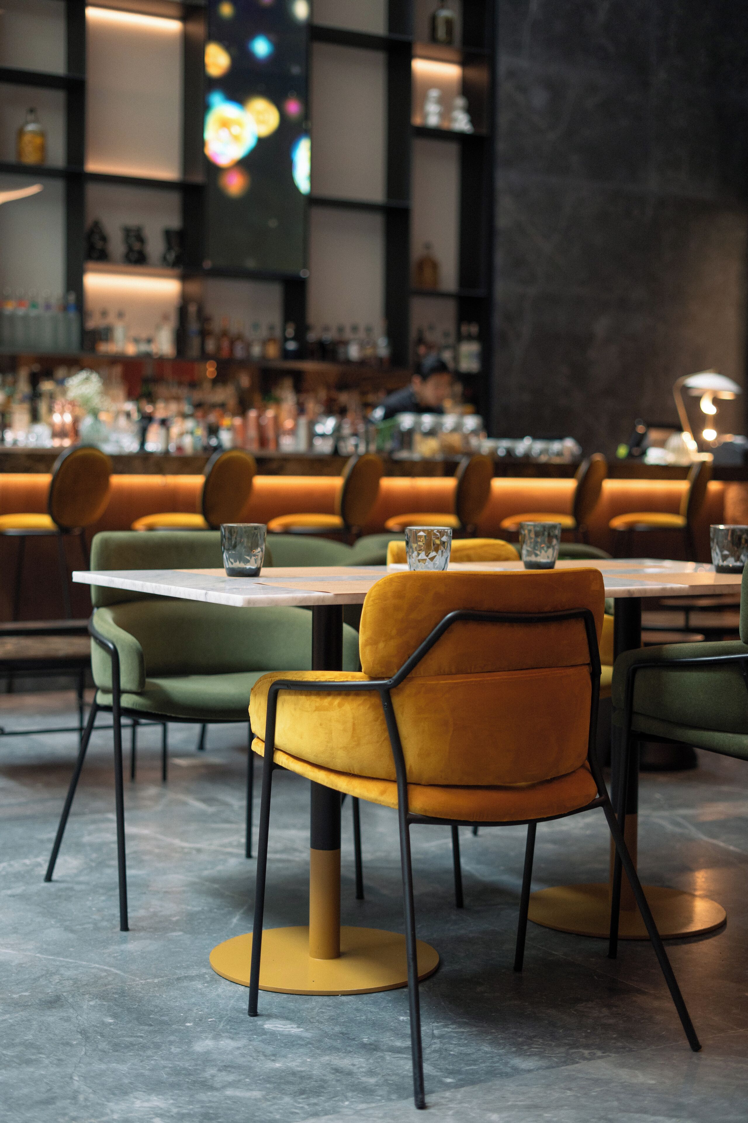 Interior de restaurante con sillas tapizadas en verde y amarillo, zona de bar con estantes llenos de botellas y un diseño moderno.
