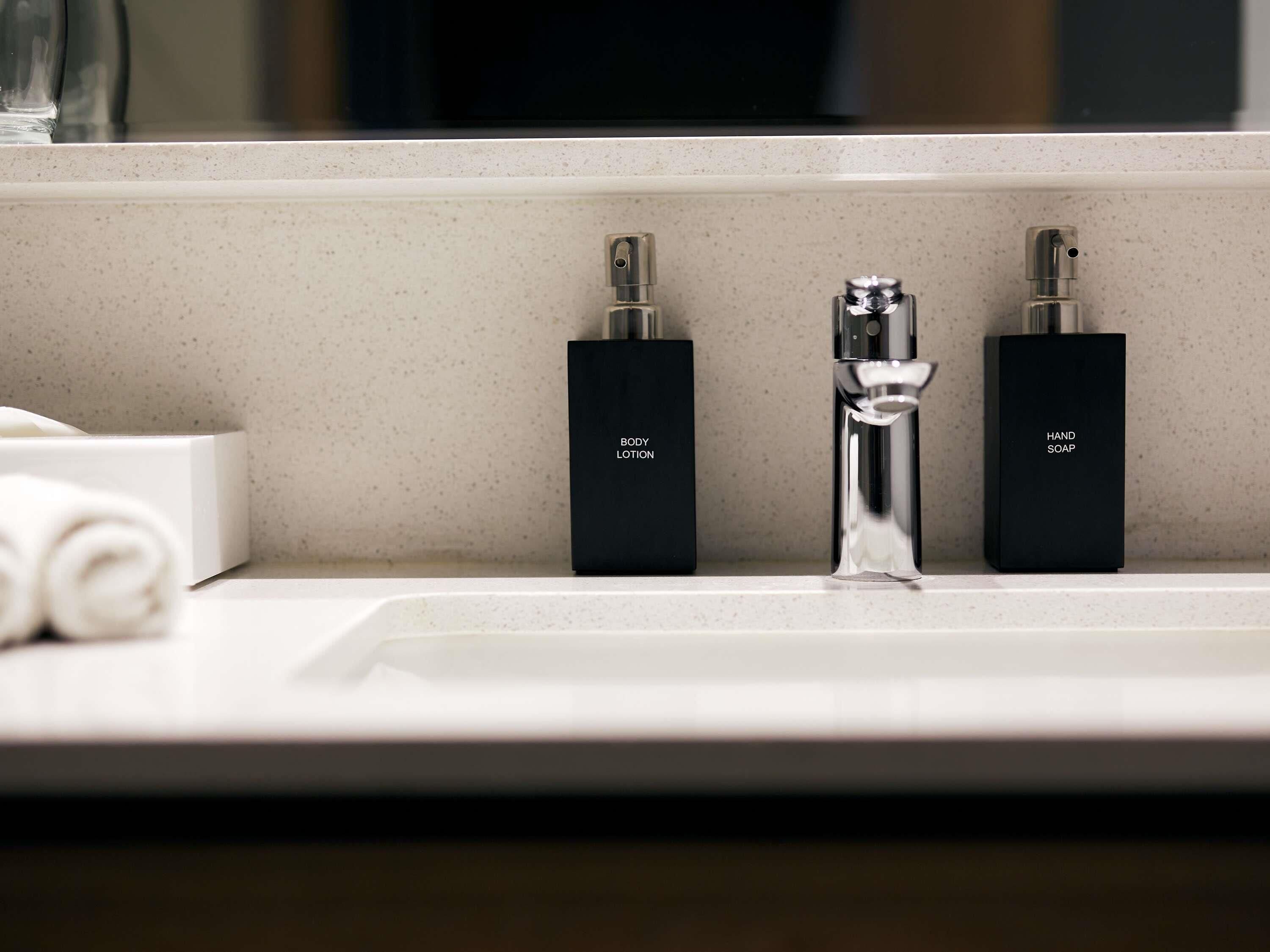 A bathroom sink with two black soap dispensers labeled "BODY LOTION" and "HAND SOAP" and a white towel on the counter.
