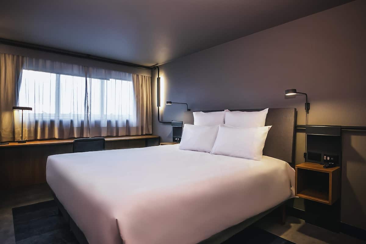 Ein Hotelzimmer mit einem großen Bett, Nachttischen, einem Schreibtisch, einem Sessel und einem Fenster mit Vorhängen.