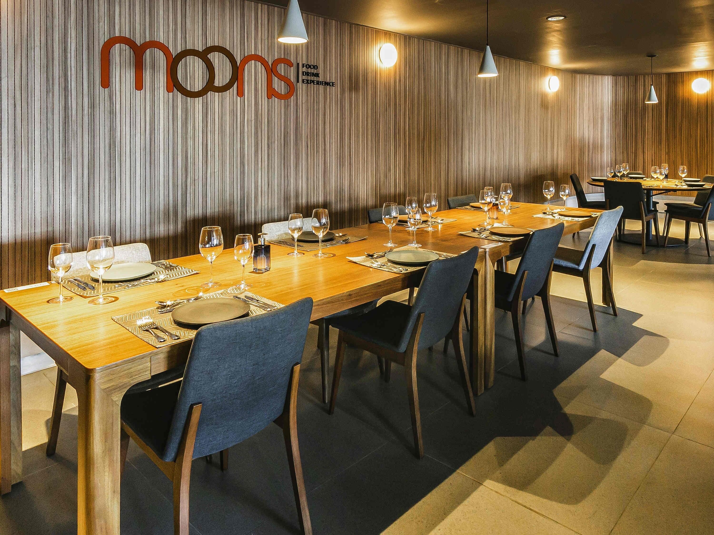 Un restaurant avec une longue table en bois, dressée pour le repas, des chaises et un mur orné de lignes verticales et d’un logo.