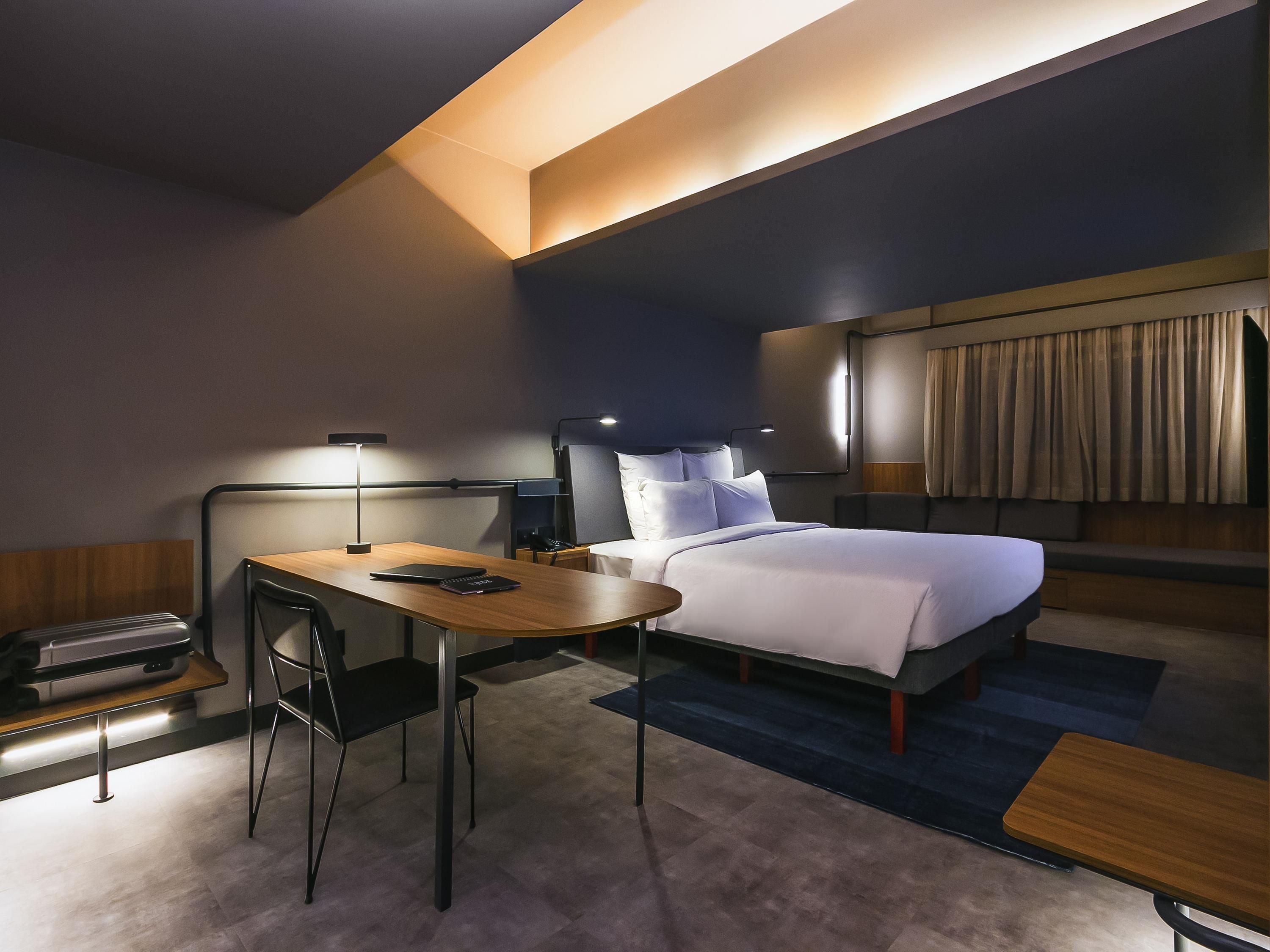 A hotel room with a bed, bedside table, desk, chair, and a desk lamp.