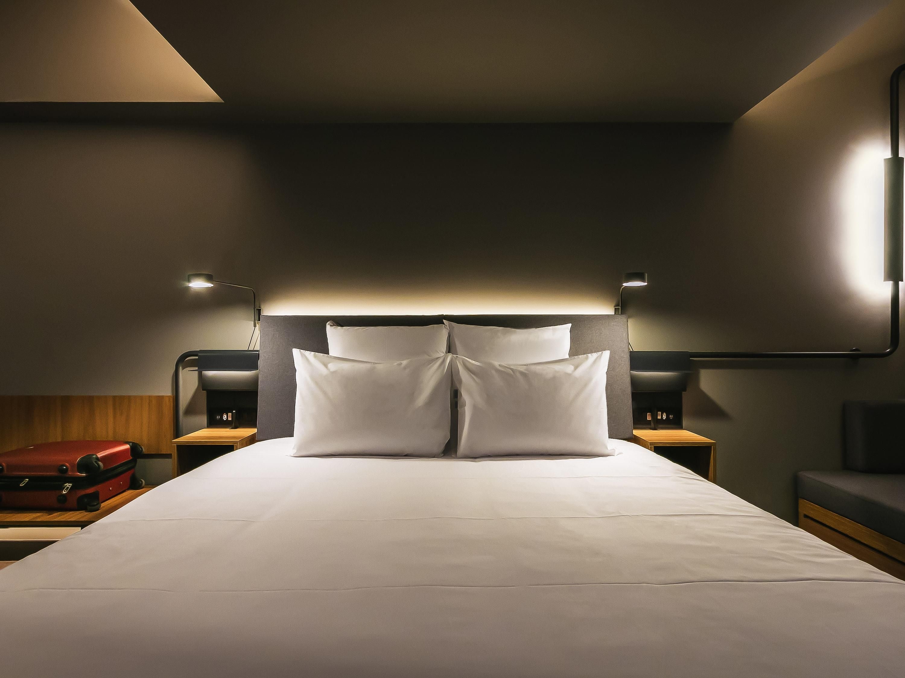 A modern hotel room with a large bed, two bedside tables, and a headboard with integrated lighting.