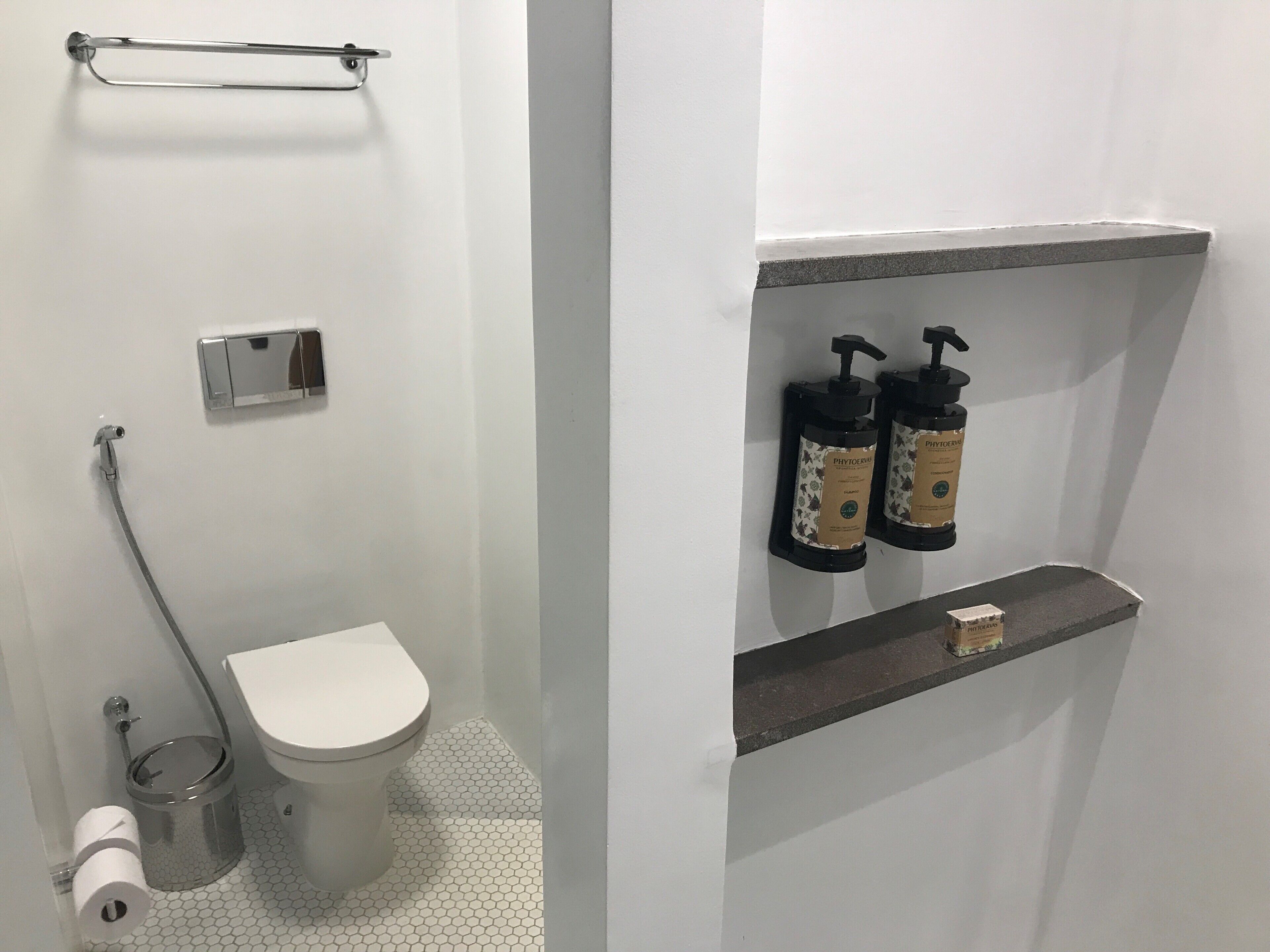 A bathroom with a toilet, a hand dryer, a trash can, and a shelf with two bottles of hand soap.
