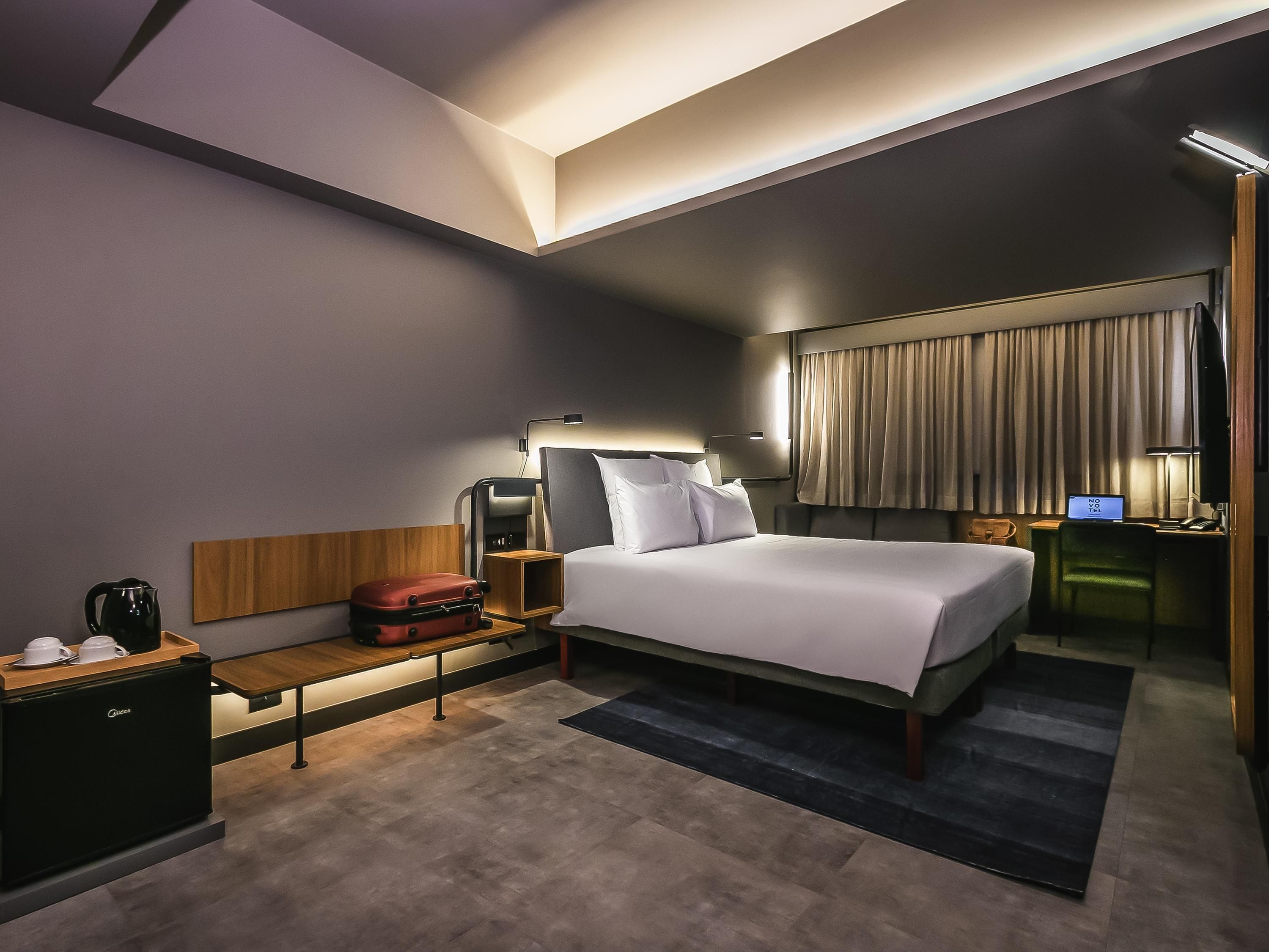 A modern hotel room with a large bed, bedside tables, a desk with a computer, and a view of the city.