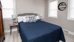 Ein Schlafzimmer mit einem Bett, zwei Nachttischen, einer Lampe, einem Wandregal und einem Fenster mit Jalousien.