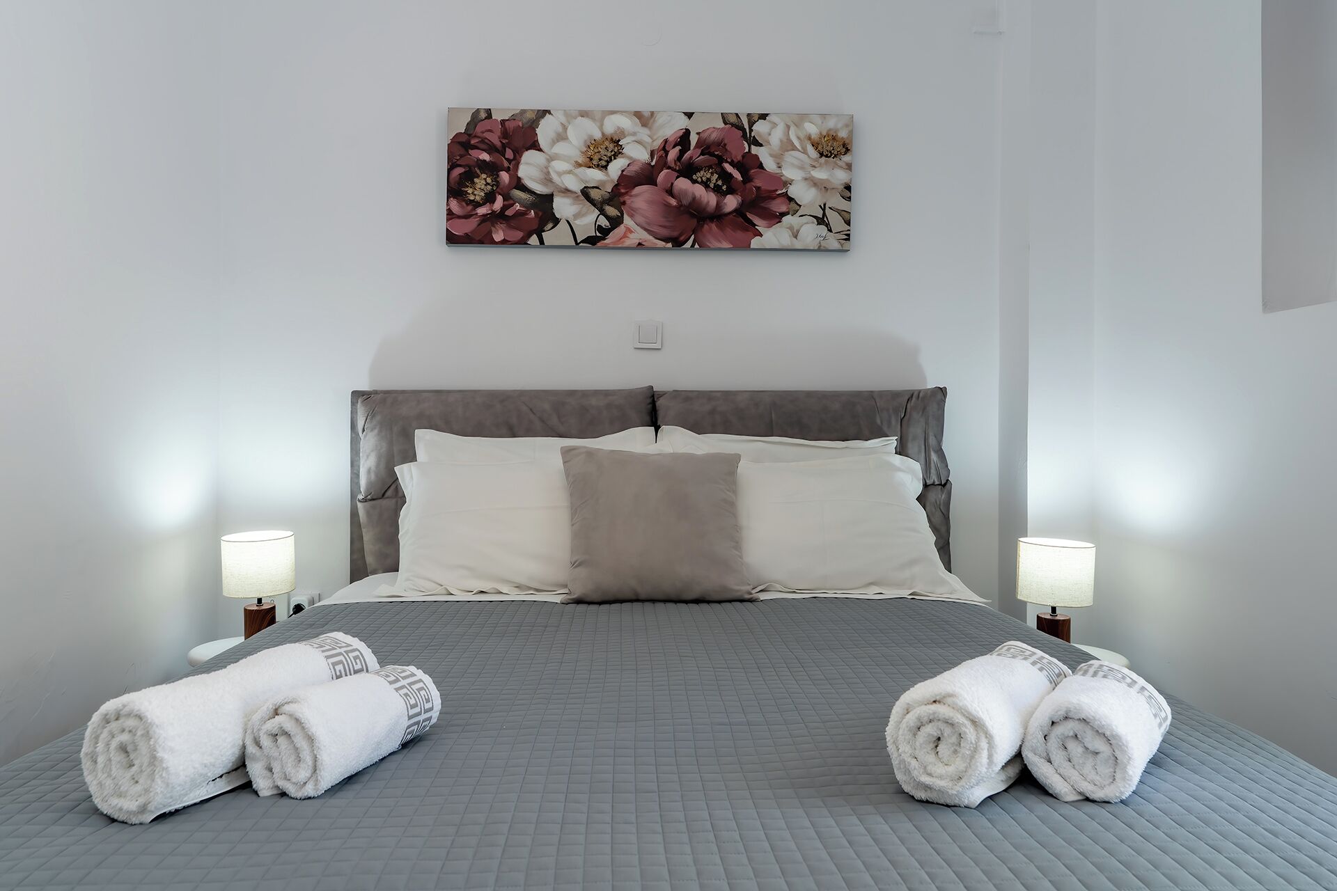 A neatly made bed with a grey quilt, two white pillows, and two rolled towels on the side.