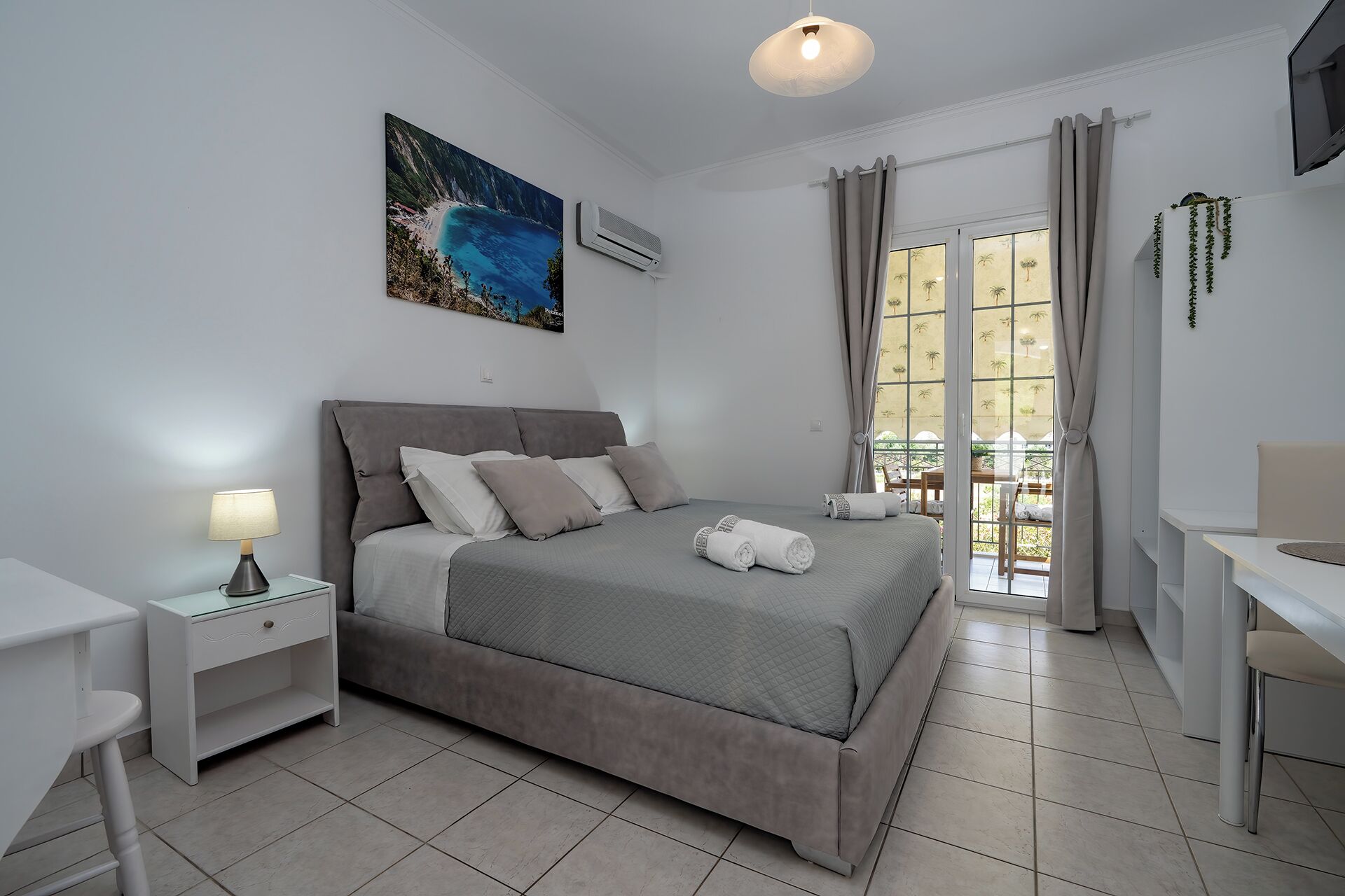 Quarto com cama, mesa de cabeceira, luminária, aparelho de ar condicionado e vista para o exterior através de uma porta de vidro.
