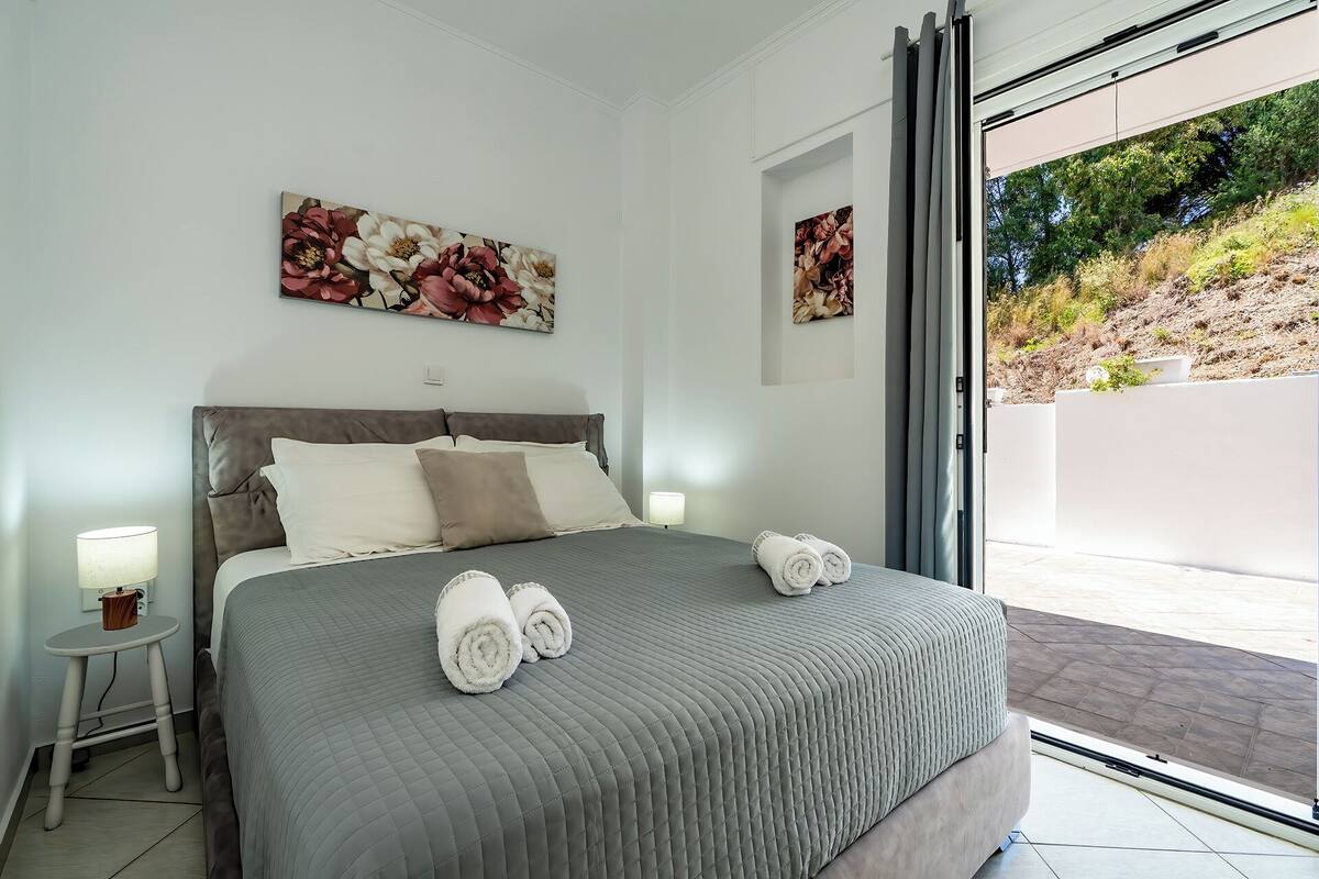A bedroom with a bed, two wall-mounted artworks, a bedside table with a lamp, and a view of a landscaped area.