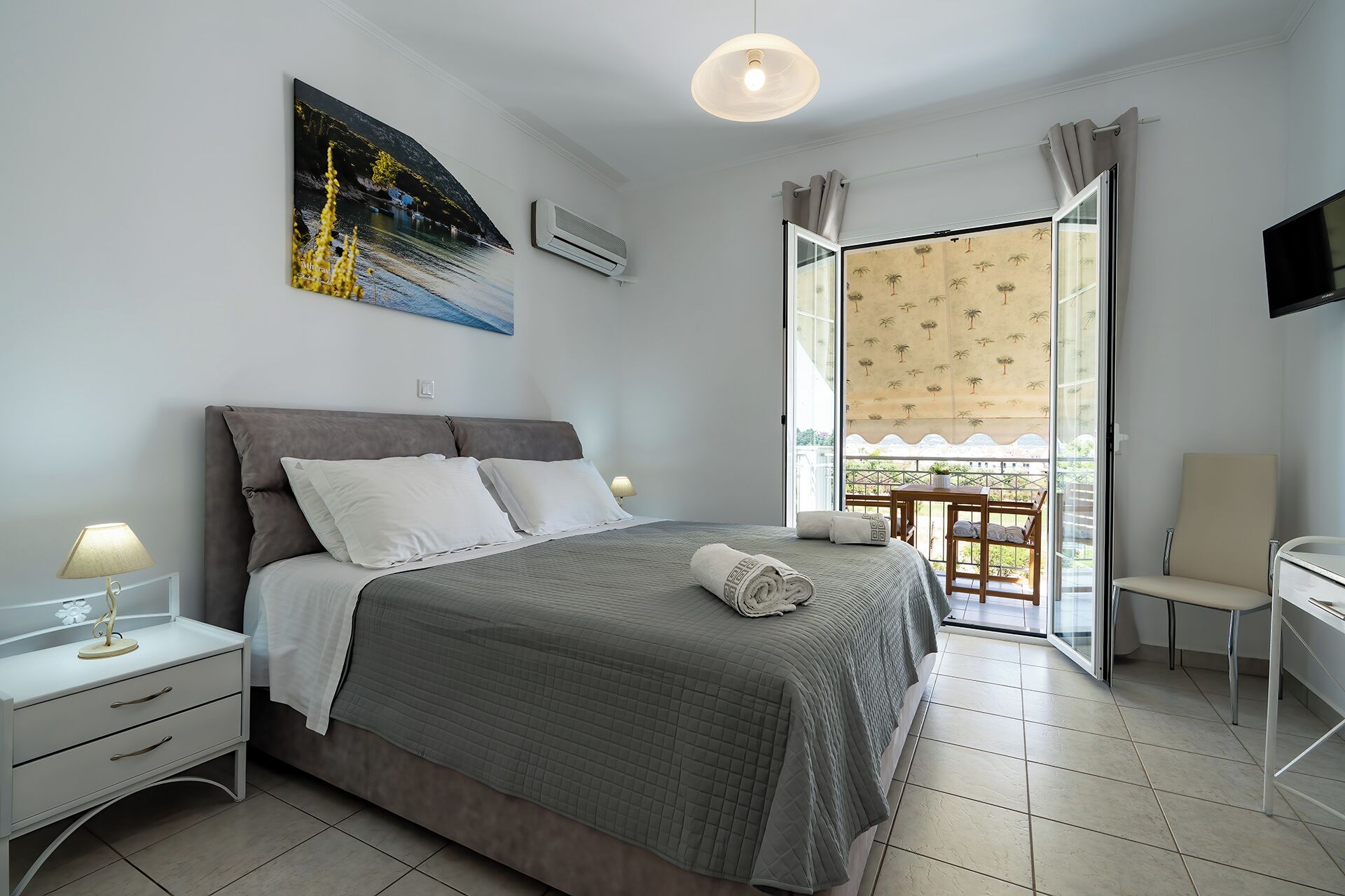 A bedroom with a bed, bedside tables, a desk, a chair, a TV, and a balcony with a view.
