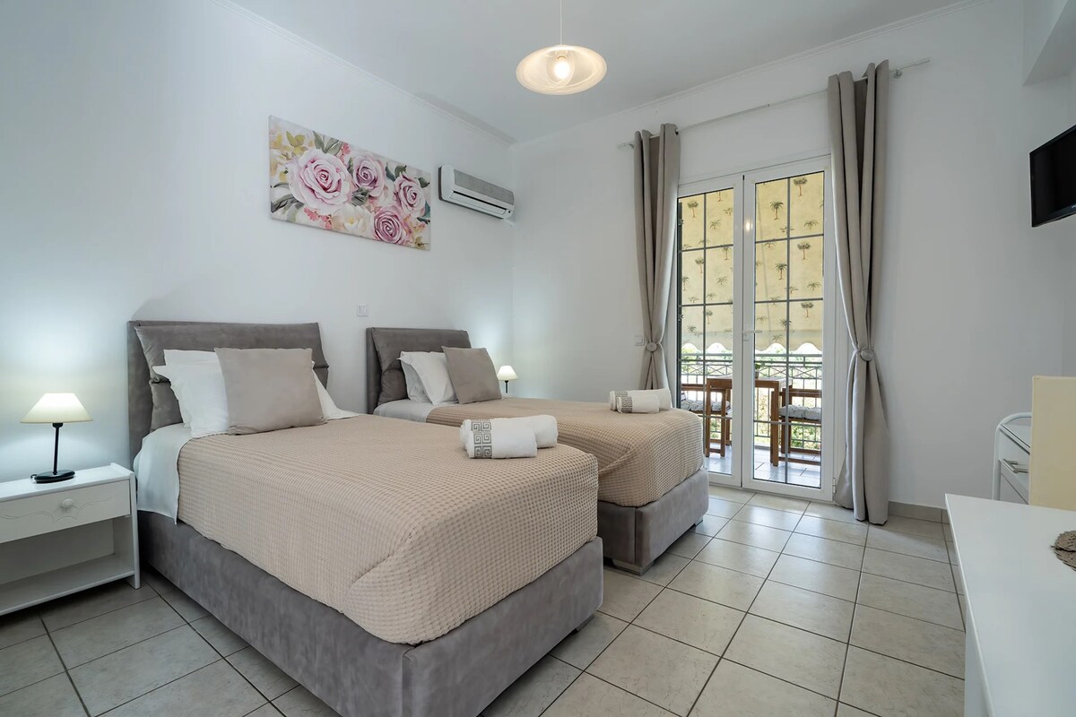 A hotel room with two beds, a nightstand with a lamp, an air conditioner, a large window with curtains, and a view of the outside.