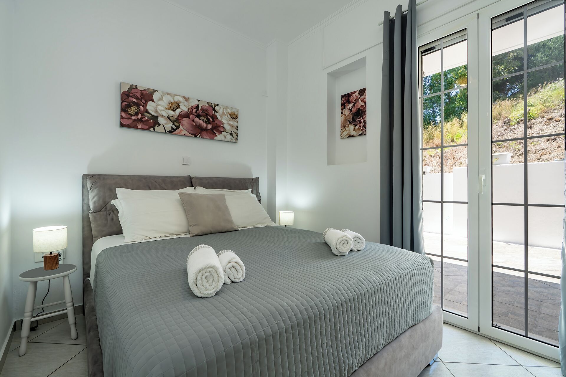 Un dormitorio con una cama, dos cuadros de flores en la pared, una mesita de noche con lámpara y vistas a través de una puerta corrediza de vidrio.