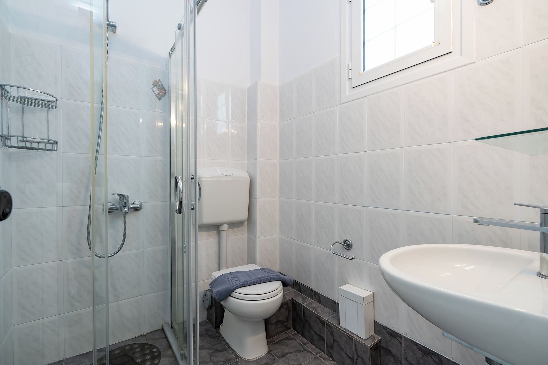Un baño con cabina de ducha de vidrio, inodoro, lavamanos y un ventanal pequeño.