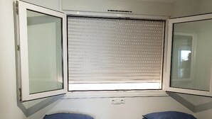 Une petite chambre avec deux lits simples, chacun doté d’un matelas et d’un oreiller bleus. Il y a une fenêtre avec des stores et un petit climatiseur placé au-dessus de la fenêtre.