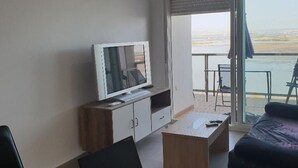 Une chambre moderne équipée d’un ordinateur portable, d’une télévision et d’un carnet sur un bureau, ainsi que d’un canapé et d’un balcon offrant une vue sur l’eau.