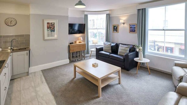A modern living room with a sofa, coffee table, and a TV. There is a kitchen area with a sink and cabinets. - Host & Stay | The Glaisdale (Whitby)
