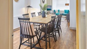 Un coin repas avec une table en bois et des chaises noires, une plante en pot, et une vue sur un salon avec des fauteuils turquoise et une fenêtre avec des stores.