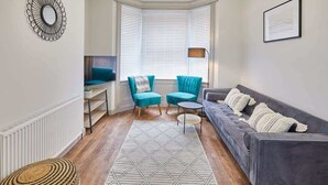 Un salon avec un canapé gris, deux fauteuils turquoise, une petite table ronde, un tapis à motifs et une baie vitrée avec des stores.