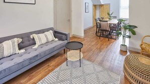 Un salon avec un canapé gris, une petite table ronde et un tapis à motifs géométriques. On y trouve également un coin repas avec une table et des chaises en bois, ainsi qu’une plante en pot.
