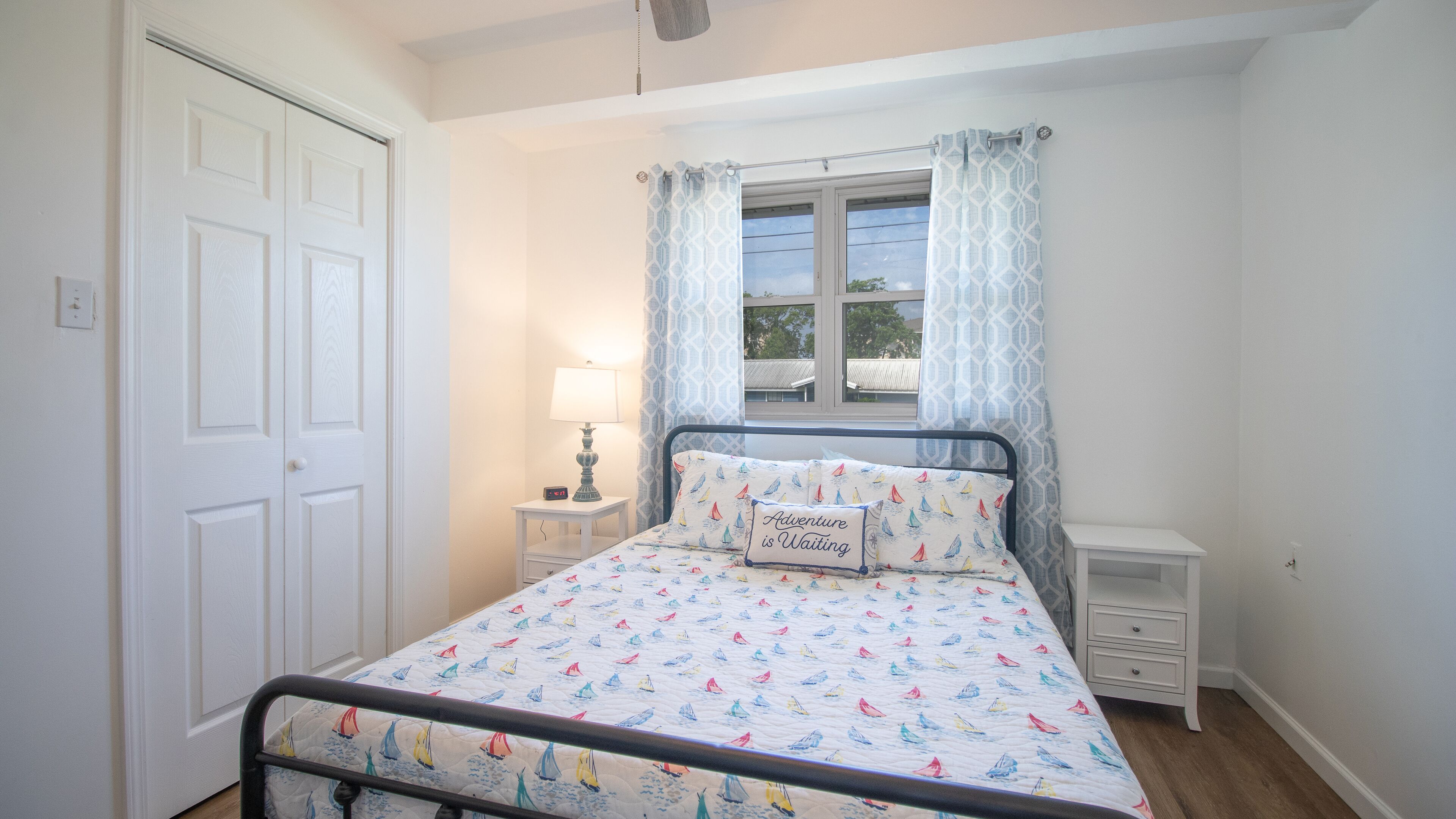 A bedroom with a bed, a nightstand, a lamp, a window with curtains, and a door.
