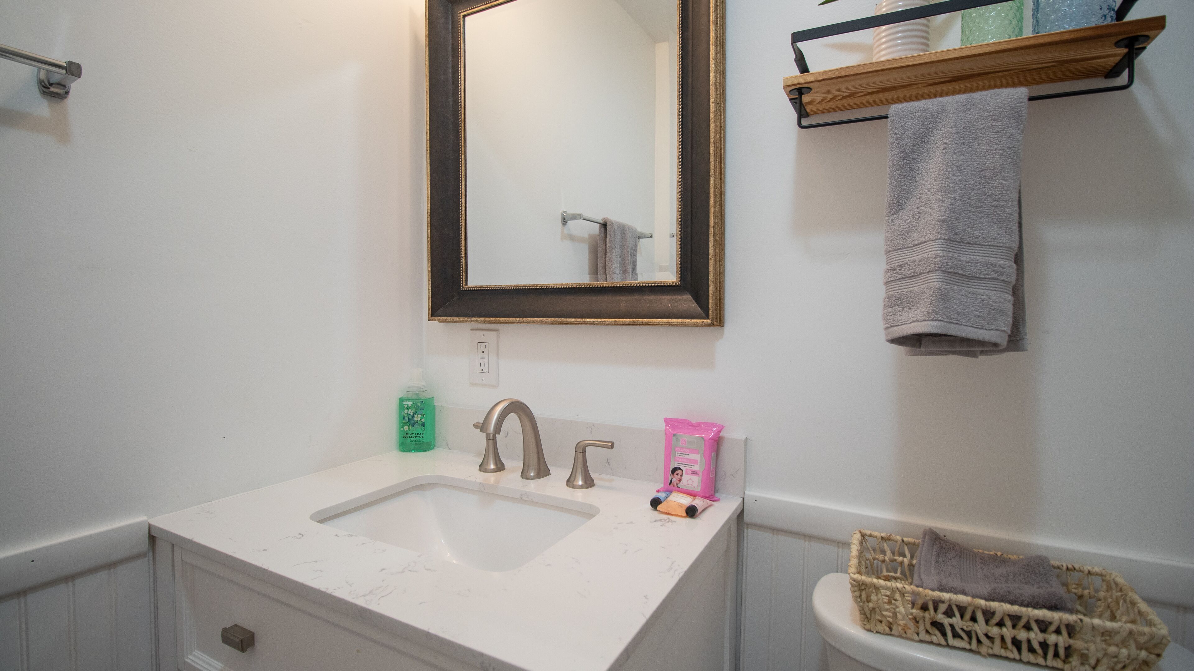 Un baño con lavamanos blanco, un espejo, toallero y una cesta.