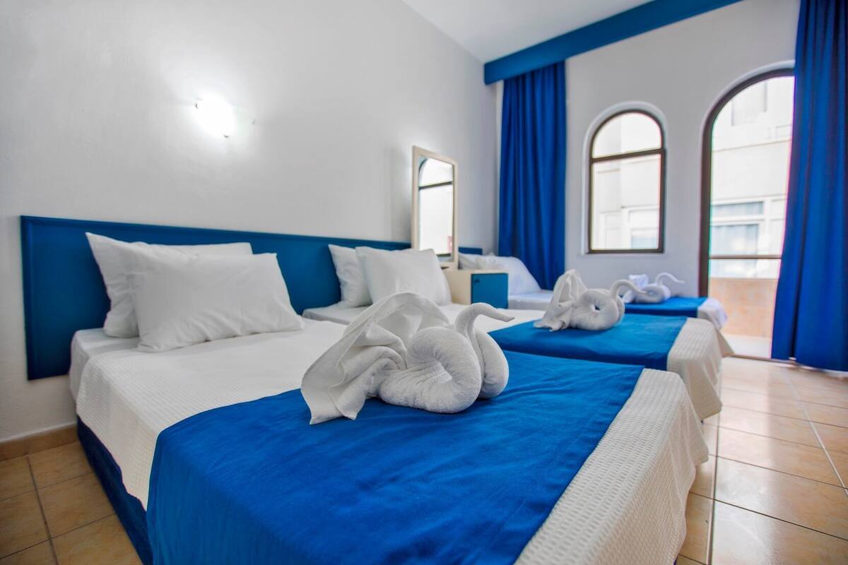 A hotel room with two beds, blue headboards, white bedding, and a view through large windows.