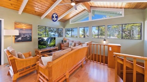 A spacious living room with a wooden ceiling, large windows with tropical views, a comfortable sofa, and a flat-screen TV. - Large Home in Waikoloa Village | 5-Bedrooms | Newly Updated with AC (Waikoloa)